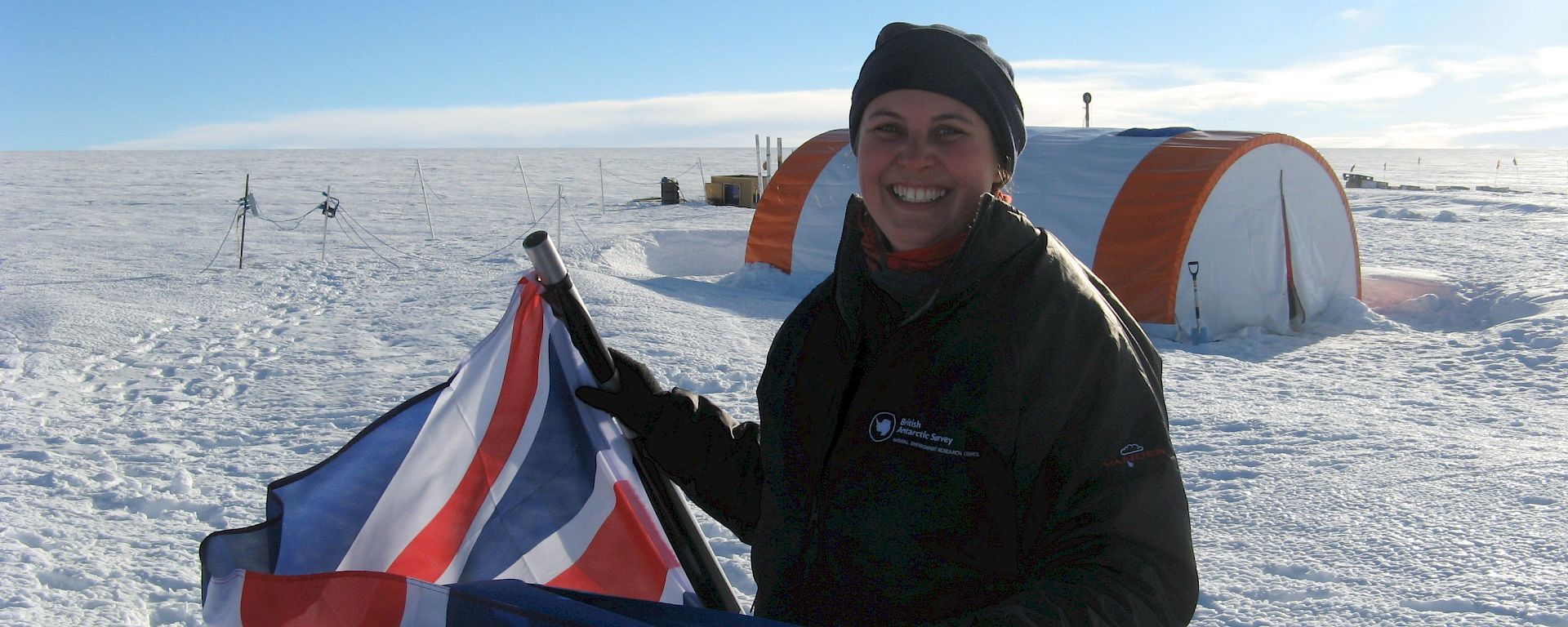 A woman holding a British flag outside cylindrical ice core drilling tent in Antarctica.