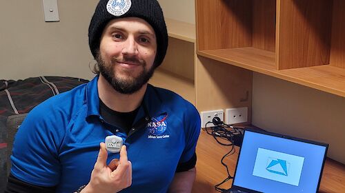 Antarctic station doctor holding a small wearable biosensor. He is seated at a desk with a laptop with one of the cognitive shape tests visible on the screen.