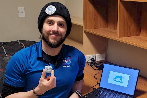 Antarctic station doctor holding a small wearable biosensor. He is seated at a desk with a laptop with one of the cognitive shape tests visible on the screen.