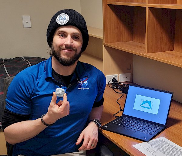 Antarctic station doctor holding a small wearable biosensor. He is seated at a desk with a laptop with one of the cognitive shape tests visible on the screen.