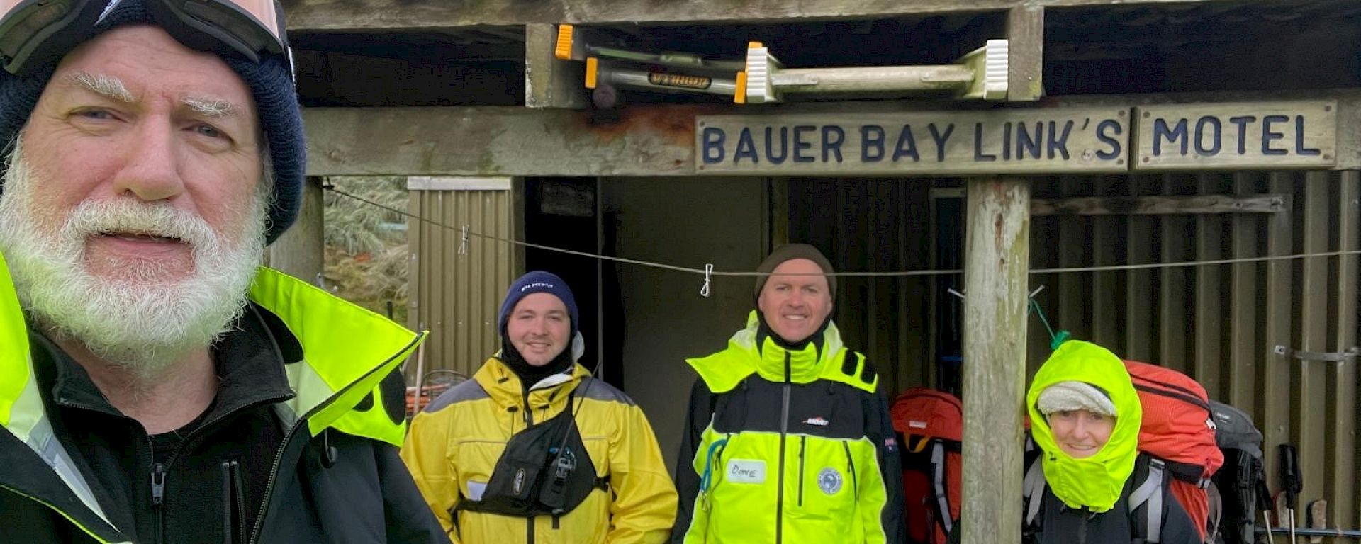 A group of four smiling people wearing bright yellow field clothing stand in front of a hut with the sign "Bauer Bay Links Motel" on it.