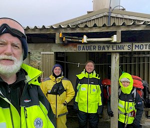 A group of four smiling people wearing bright yellow field clothing stand in front of a hut with the sign "Bauer Bay Links Motel" on it.