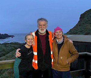 Three smiling people with their arms around one another's shoulders pose for a farewell shot with the ship just visible in the background.