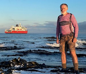 A man wearing a checked shirt and lederhosen stands on the snowy seashore with a red icebreaker in the ocean behind him.