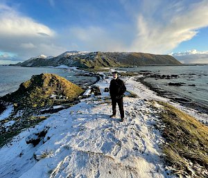 A man in black stands atop a snow-covered hill with an isthmus and buildings behind him below and a plateau in the distance.