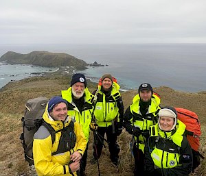 A group of five people wearing bright yellow hiking gear stand on top of a grassy hill with an isthmus and headland in the background surrounded by grey ocean and cloudy skies.
