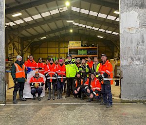 A group of people mostly wearing bright orange high vis clothing stand in a group pose surrounding a tall man with a beard. The group is standing in the opening to a large industrial shed.