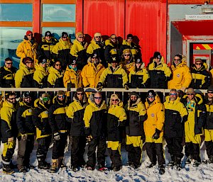 The Casey Station Winter 2025 team all dressed in yellow and black AAD clothing standing outside the Red Shed