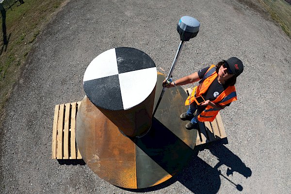 A surveyor stands beside a metal reflector shaped like a top hat, with a black and white checkerboard pattern on its round top.