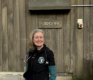 A smiling woman with a stethoscope around her neck stands in front of a 'Surgery' sign on a wooden wall.