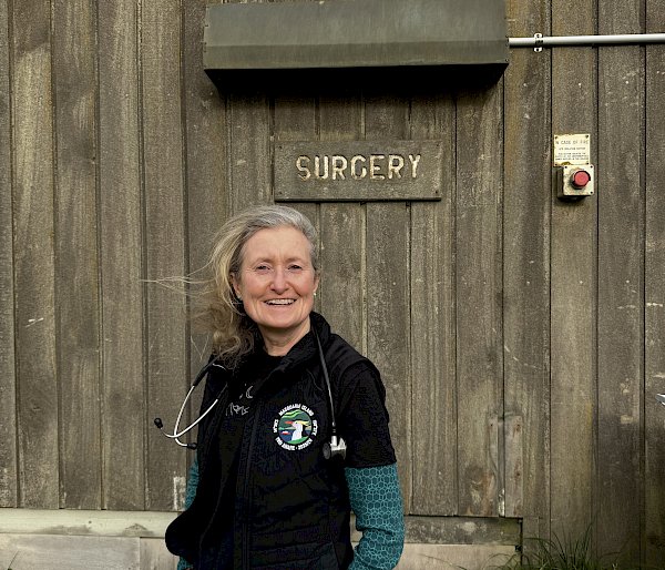 A smiling woman with a stethoscope around her neck stands in front of a 'Surgery' sign on a wooden wall.