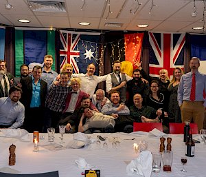 A group photo of the Mawson expeditioners arranged behind a large table that is covered in the remnants of a festive dinner