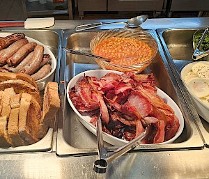 A bain marie filled with backon, sausages, bread, baked beans and eggs