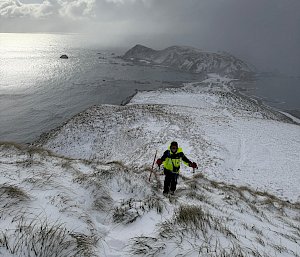 A hiker with walking poles stands at the top of a snow-covered hill. In the background some distance away is a snow-covered isthmus surrounded by ocean.