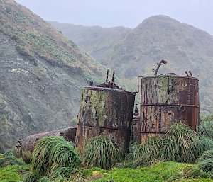 Two old rusty cylindrical digesters sitting amidst green tussock grass and surrounded by green and rocky mountainous terrain.