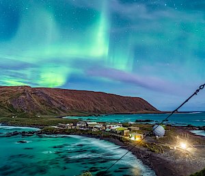 A dazzling green and purple aurora lights up the night sky above a mountain range. There is an ocean and isthmus in the foreground.