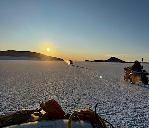 Three quad bike riders on a flat expanse of ice, driving towards a sunset
