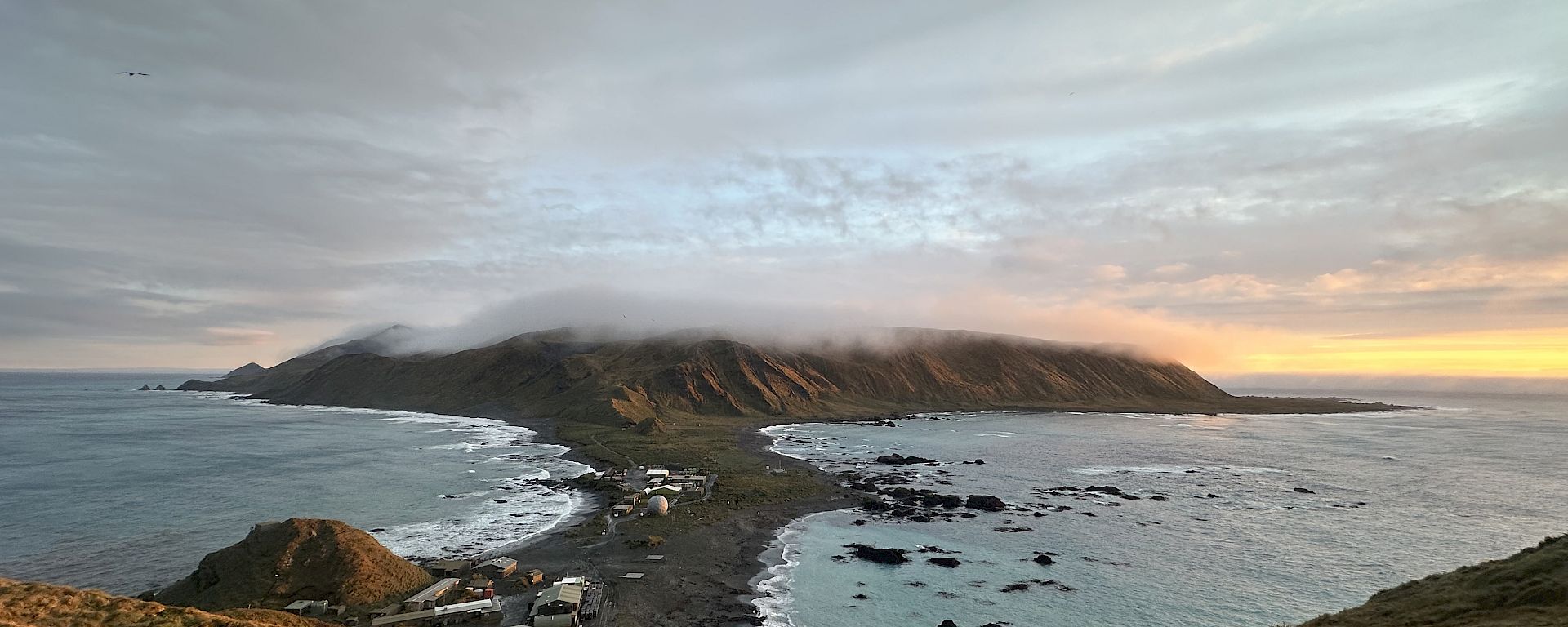 A panoramic view of the Macquarie Island Isthmus viewed from a high plateau. There is cloud hovering over a distant headland and golden skies as the sun sets in the distance.