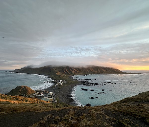 A panoramic view of the Macquarie Island Isthmus viewed from a high plateau. There is cloud hovering over a distant headland and golden skies as the sun sets in the distance.