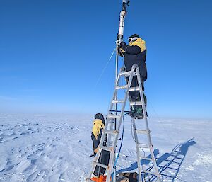 Two expeditioners working on a mast and equipment up a ladder on the snow