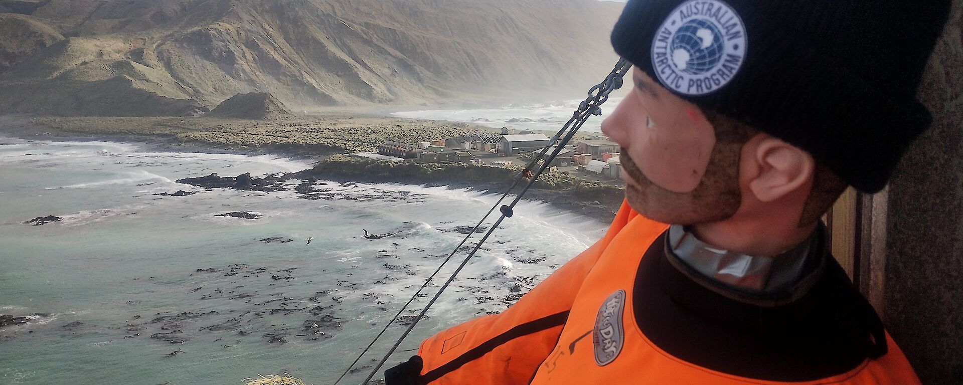 A life-sized dummy wearing an orange suit and  black beanie with an Australian Antarctic Division logo on it, sits on a balcony overlooking the Macquarie Island isthmus and plateau.