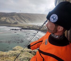 A life-sized dummy wearing an orange suit and  black beanie with an Australian Antarctic Division logo on it, sits on a balcony overlooking the Macquarie Island isthmus and plateau.