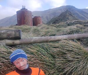 A life-sized dummy wearing an orange suit sports a hand knitted striped grey and blue beanie. He is leaning against green tussock grass and there is a copper log pine fence and two rusted steel cylinders behind him.
