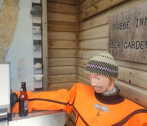 A life-sized dummy wearing an orange suit wears a traditional Pangnirtung or pang hat from Baffin Island. It is beige, green, purple and black. The dummy sits against a wooden wall holding an empty beer bottle. There is a sign on the wall saying Crabbe Inn Beer Garden