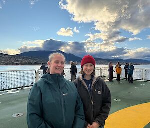 Two women standing together on the helicopter deck of a ship with a city behind them