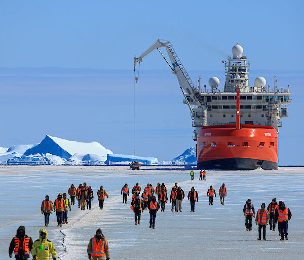 A red and white ship surrounded by ice with people walking across the ice towards it