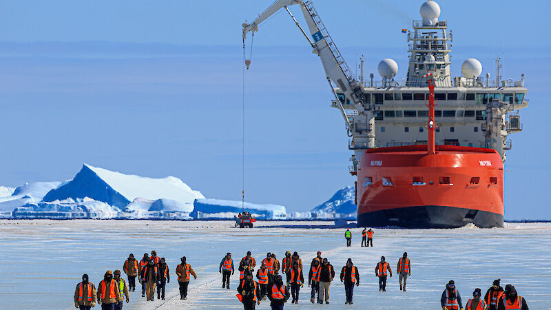 A red and white ship surrounded by ice with people walking across the ice towards it
