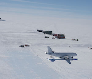 An aircraft, helicopter and oversnow vehicles near a small number of buildings on a flat, white expanse of ice.