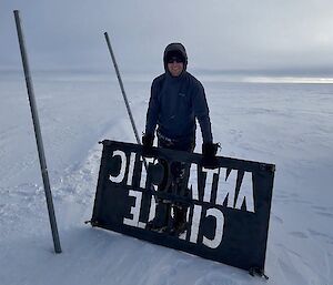 The old Antarctic Circle sign gets removed by an expeditioner standing next to the poles holding the sign standing on the ice