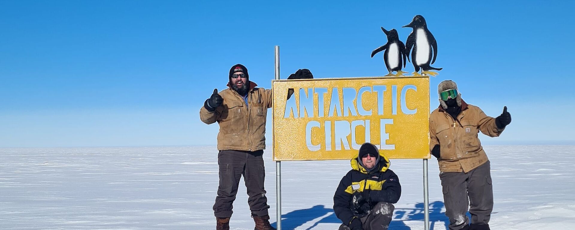 A three-person team standing around the Antarctic Circle sign