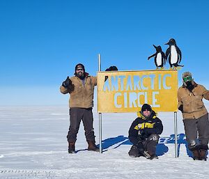 A three-person team standing around the Antarctic Circle sign