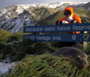 A man stands behind a sign that reads, "Macquarie Island Nature Reserve, World Heritage Area". There are snow covered hills in the background.