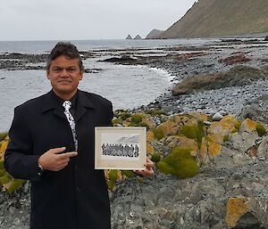 A man, standing on a rocky seashore, wearing a suit and tie holds an old black and white photograph of his father.