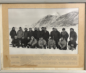 A black and white photograph of 20 men - 12 standing in the rear and 8 crouched in the front - in a snow-covered shoreline in front of mountains
