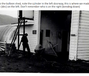 A black and white photo of two people outside a balloon shed. One is crouched down.