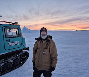 Dominik beside a hagglund on the snow, with a sunset on the horizon