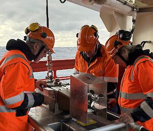 Three people in hi vis and helmets stand on a ship's deck with a metal box
