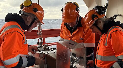 Three people in hi vis and helmets stand on a ship's deck with a metal box