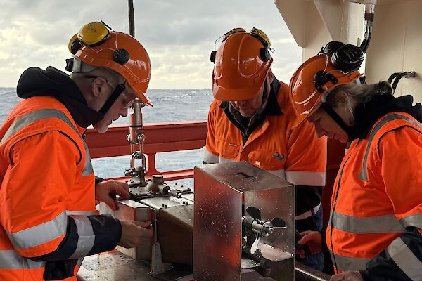 Three people in hi vis and helmets stand on a ship's deck with a metal box