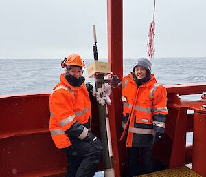Two women in hi vis stand on a ship's deck with a long science instrument
