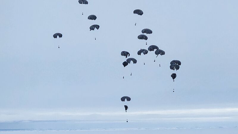 15 parachutes with bundles of cargo attached, fall through the sky above Antarctic sea ice.