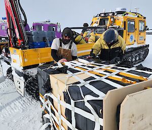 Two men standing beside a large box of cargo wrapped in a net. A hagglund carrying a small crane is behind them.