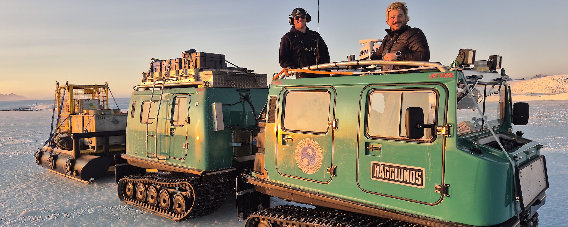 Two people standing on a twin cab Hagglund vehicle that is towing some equipment.