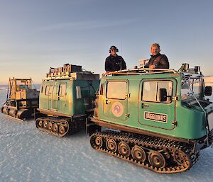 Two people standing on a twin cab Hagglund vehicle that is towing some equipment.