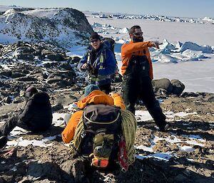 Four people sitting and standing on snow-covered rock, with a view of icebergs in the distance