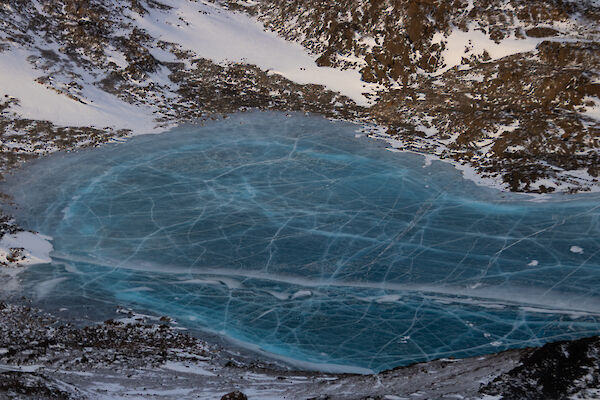 A blue frozen lake, it's surface covered in cracks, viewed from the air